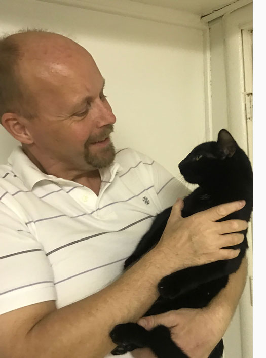 Paul is smiling and looking at a black cat that his is holding.  The cat is leaning back and looking at him.
